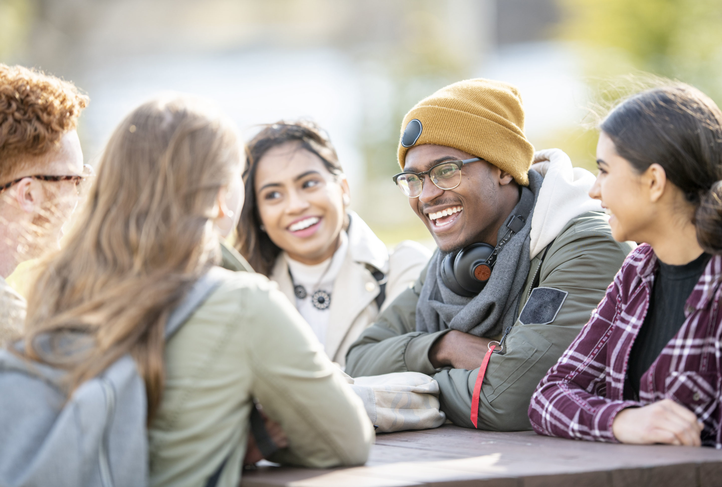 Photo: Five young adults are sitting at a picnic table outside on a fall day. They are smiling and chatting.