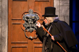 Photo of Scrooge examining metal knocker at enormous wooden door.
