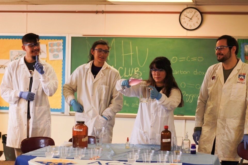 Four chemistry students perform an experiment at a desk in front of a classroom.