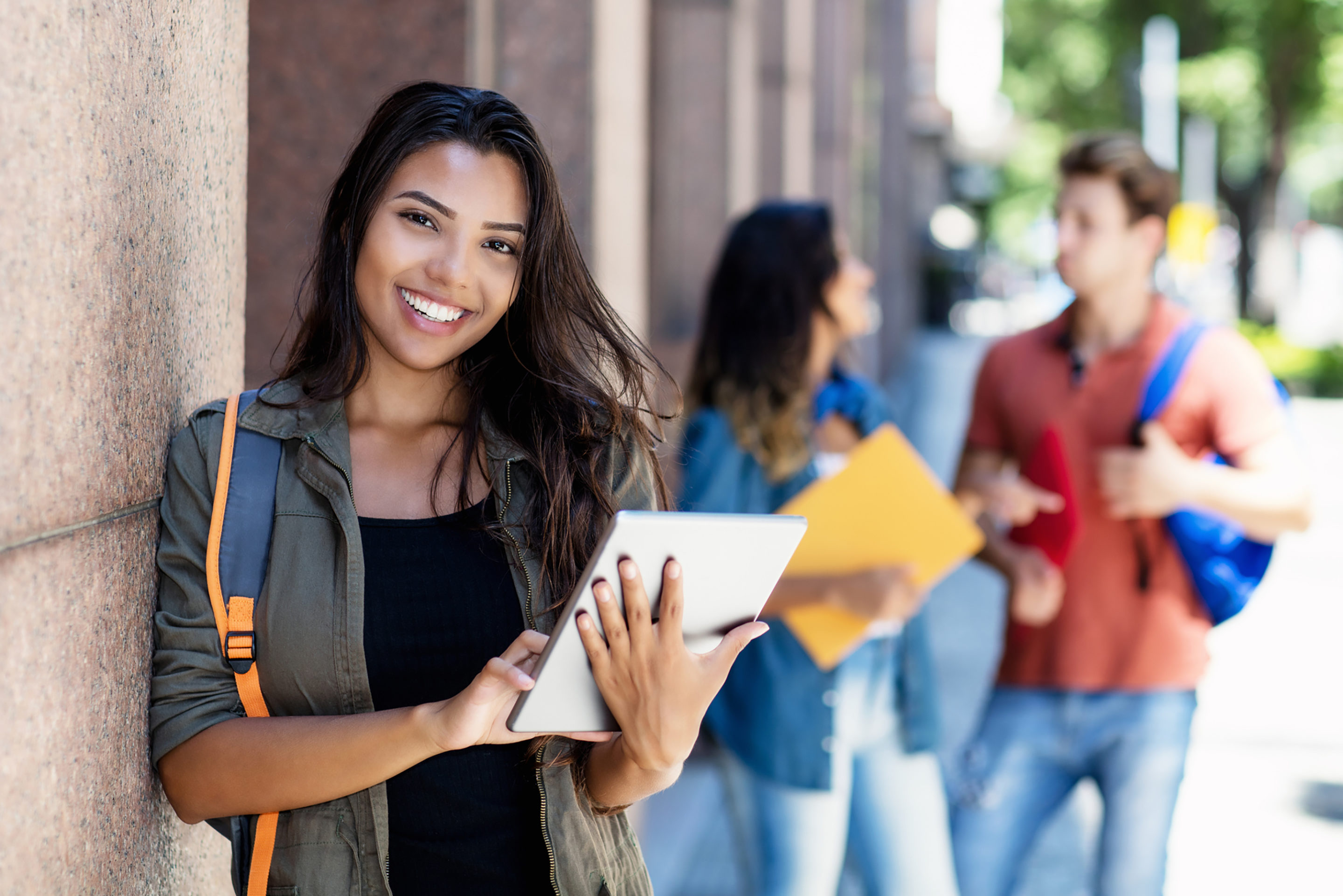 Laughing Hispanic student with tablet computer and friends in background in front of university building outdoor in summer in city