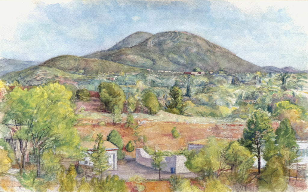 Northern New Mexico landscape with village and trees and red earth in foreground and mountains rising in background.