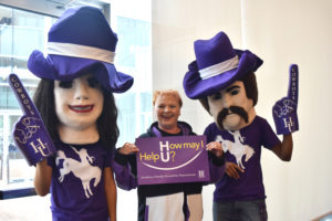A NMHU employee poses with two people with the school mascot puppet heads on. The employee holds a sign that reads: "How may I help U?"