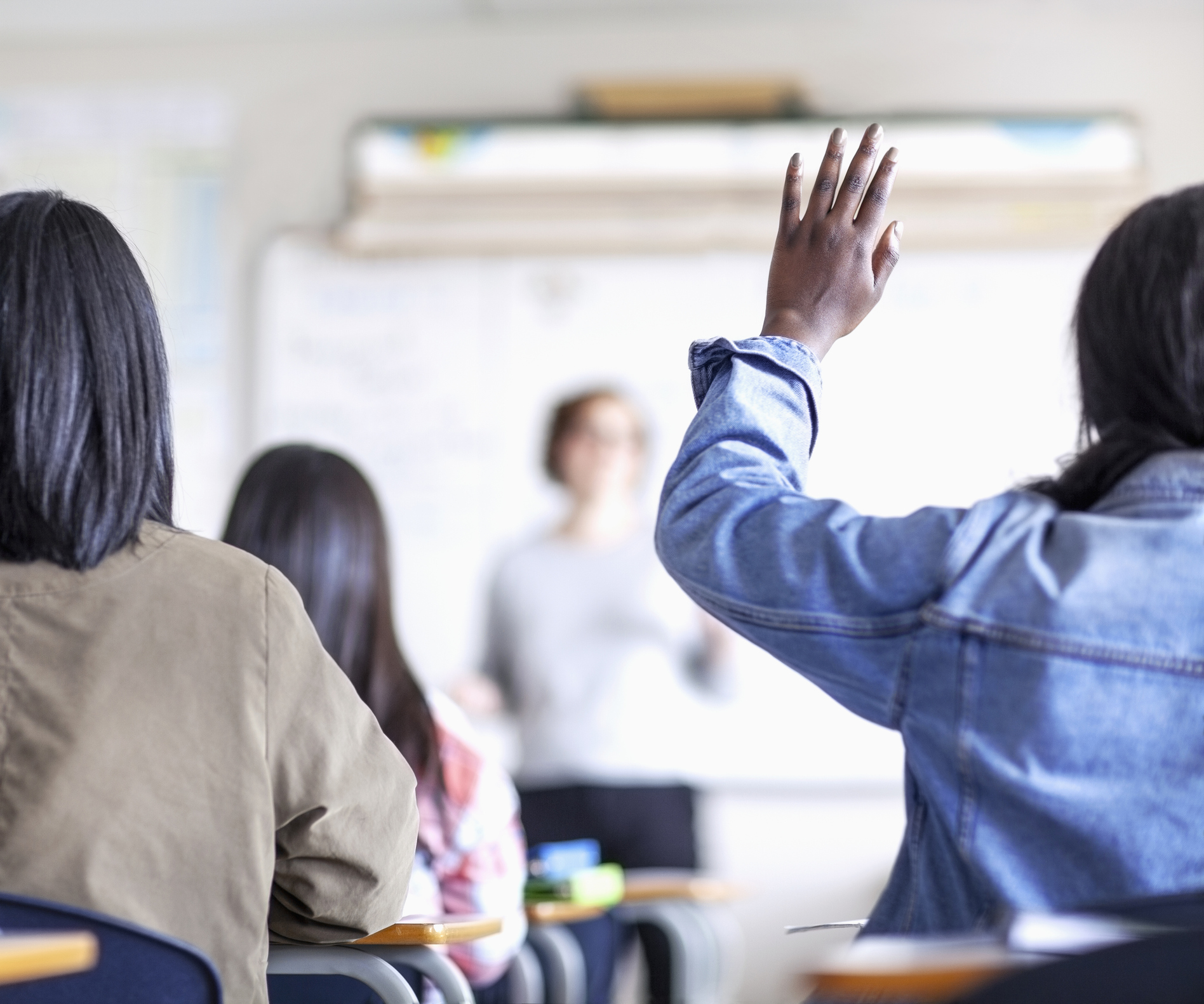 Rear view of female student asking while raising hand. Teenage girls are learning at high school. Teacher is explaining in classroom.