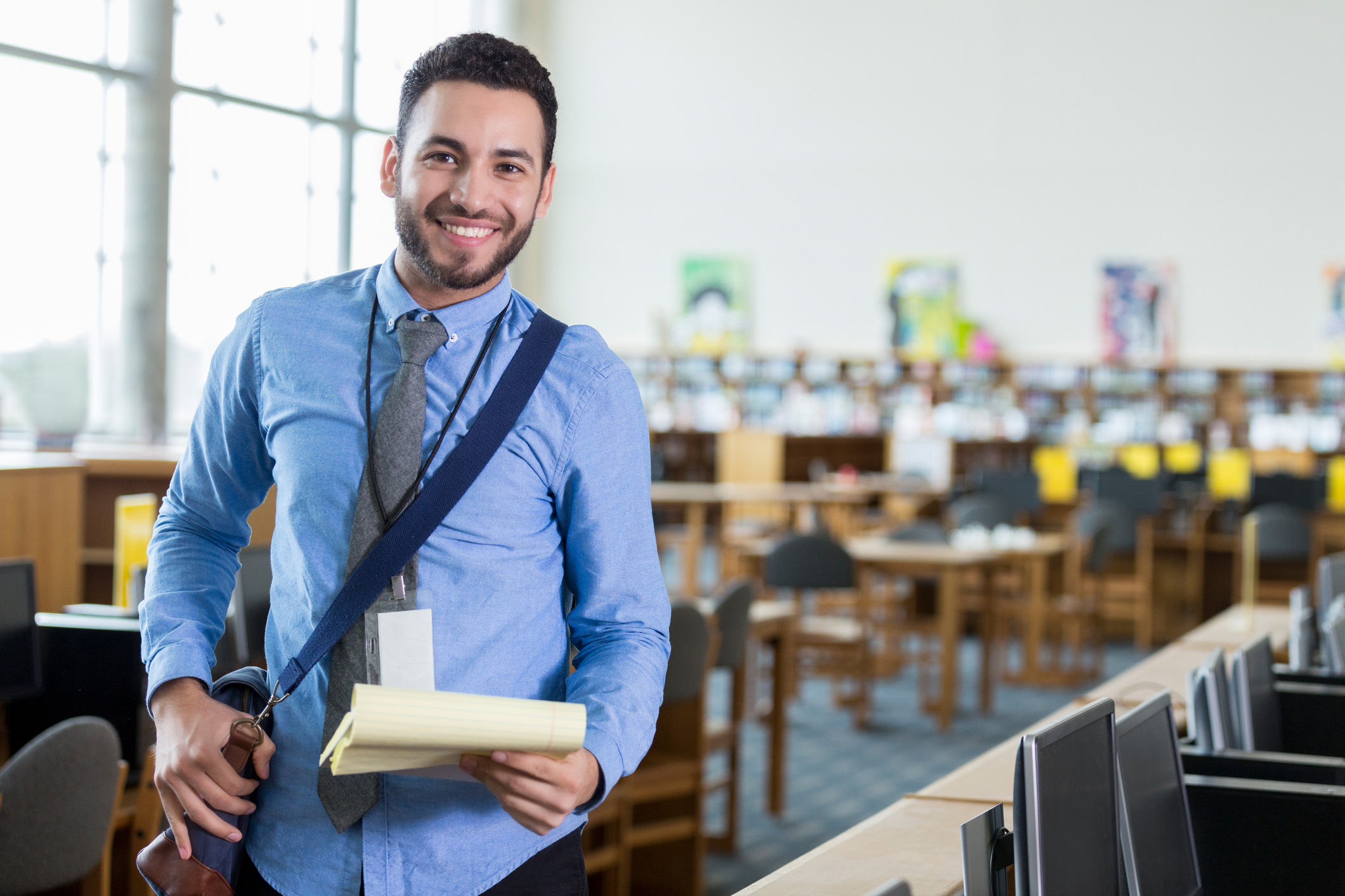 A young male Hispanic school librarian stands proudly in his library with satchel and note pad ready. He smiles for the camera.