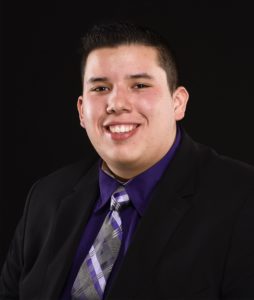 A young man smiles for the camera. He wears a black shirt and purple tie. 