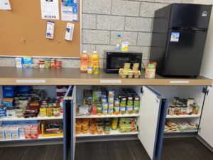 Photo of non-perishable foods on shelves, with a microwave and a small fridge.
