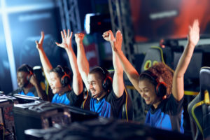 A row of smiling students sit in front of computers with their hands up, celebrating a video game victory