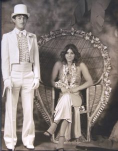 Two students from Robertson High School pose for a prom photo in 1981. The young woman has feathered hair and sits in a wicker peacock chair. The young man is wearing a white tux with a top hat and cane. 