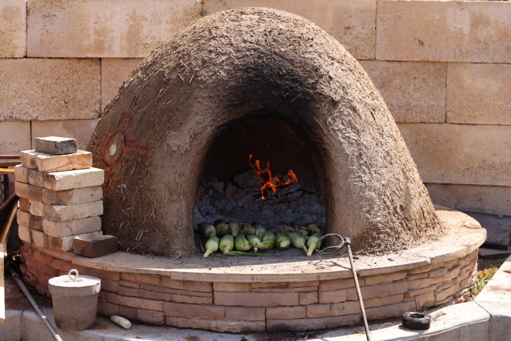A horno with a fire lit inside and corn roasting at the entry of the fire.