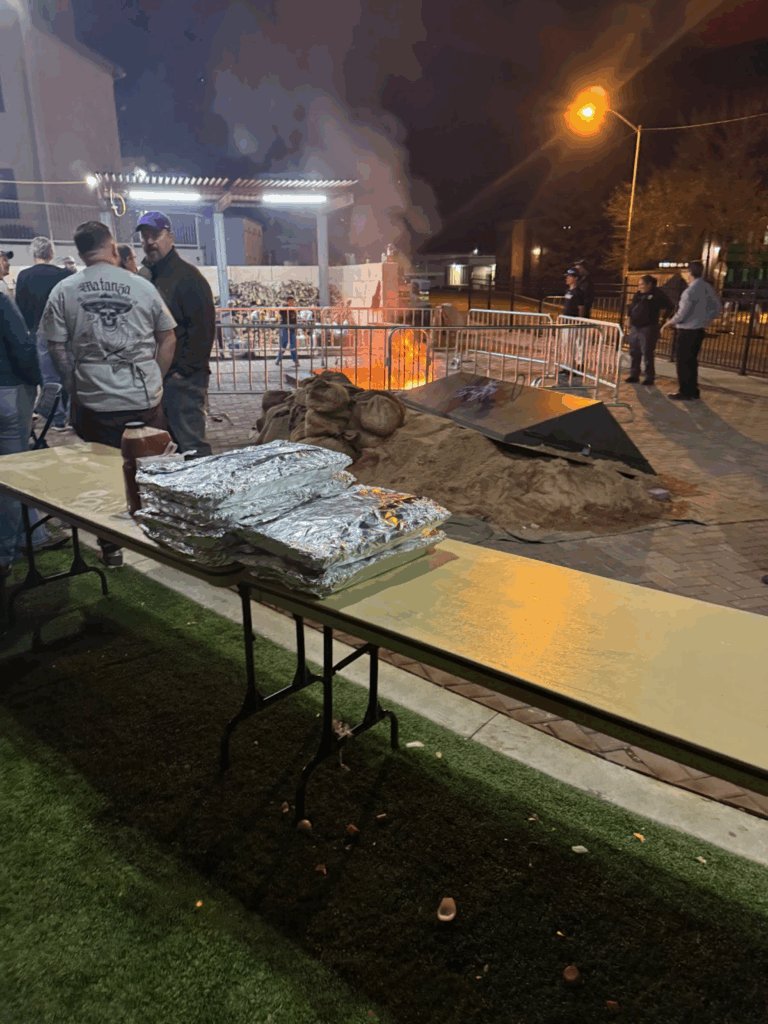 A table with foil-wrapped trays ready to be cooked, a bonfire in the firepit can be seen in the background.
