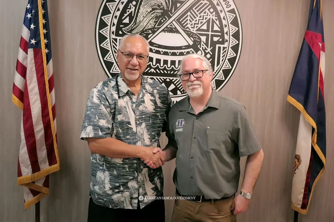 Representatives from NMHU and American Samoa shaking hands in front of the Seal for American Samoa.