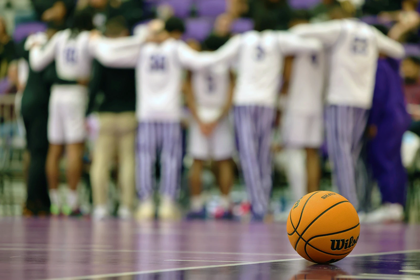 mbb-image-three Image of a basketball on the court at NMHU's John A. Wilson Center. The NMHU men's team can be seen in a huddle in the background.